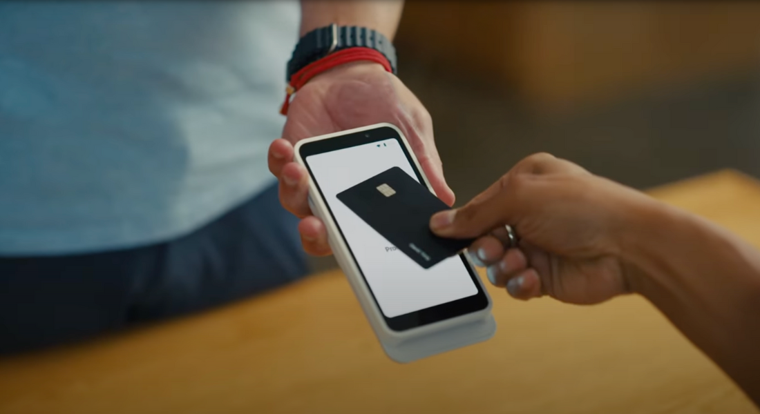 A person making a contactless payment by holding a black credit card near a handheld payment terminal or smartphone held by another person.
